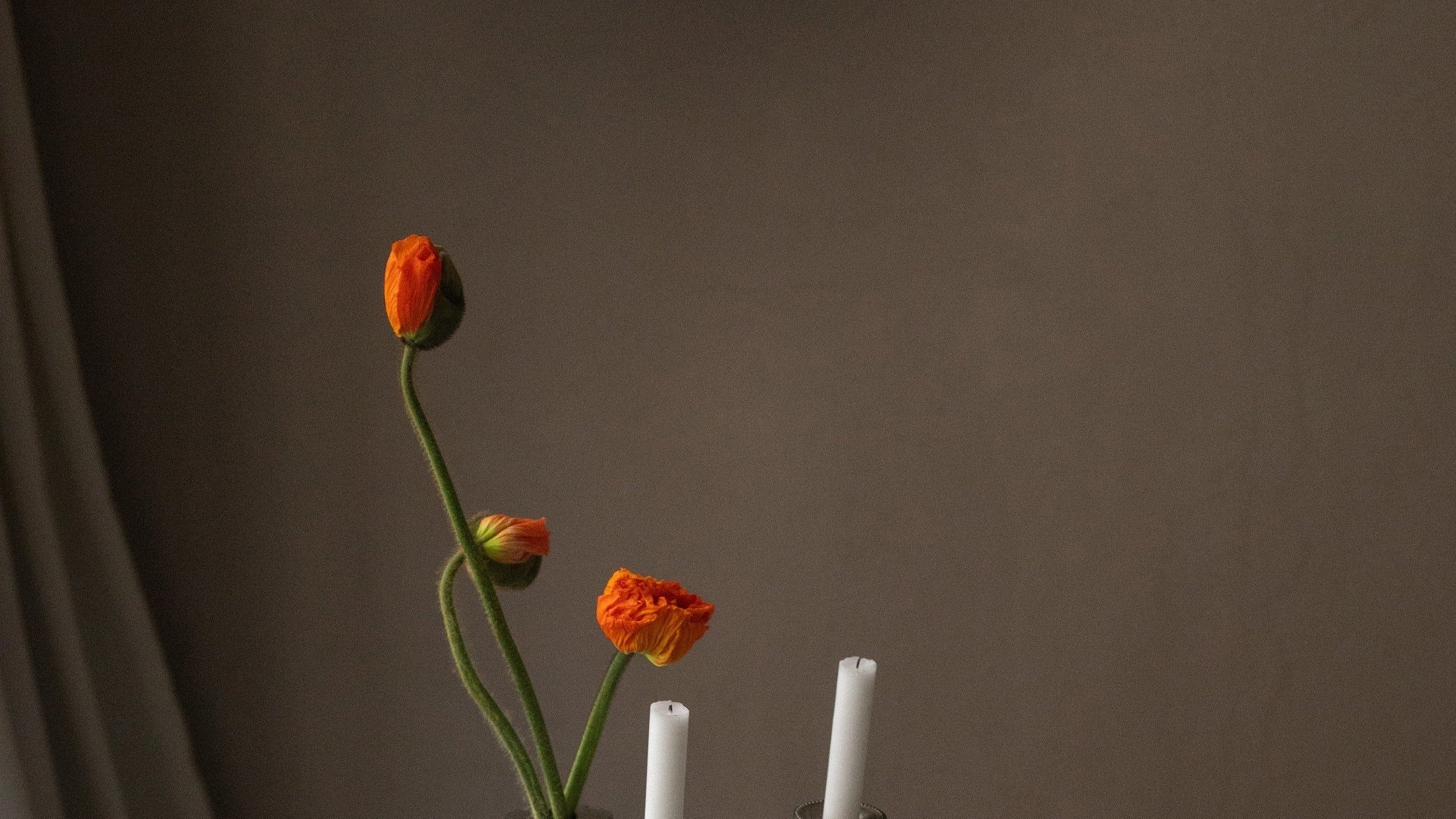Orange flowers in a vase with two white candles on a dark surface against a brown background