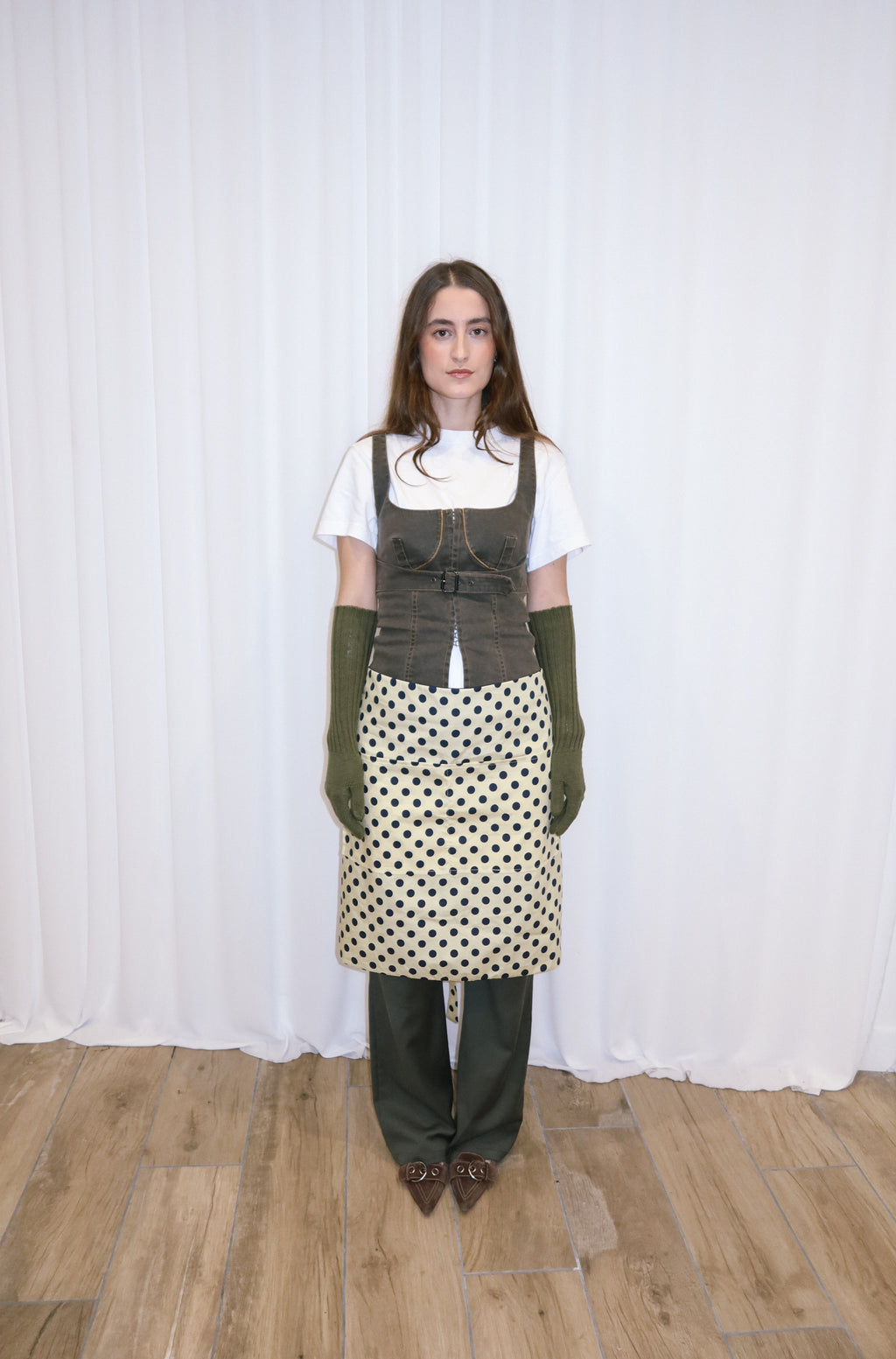 Person wearing a white shirt, polka dot apron, and green gloves against a white curtain background.
