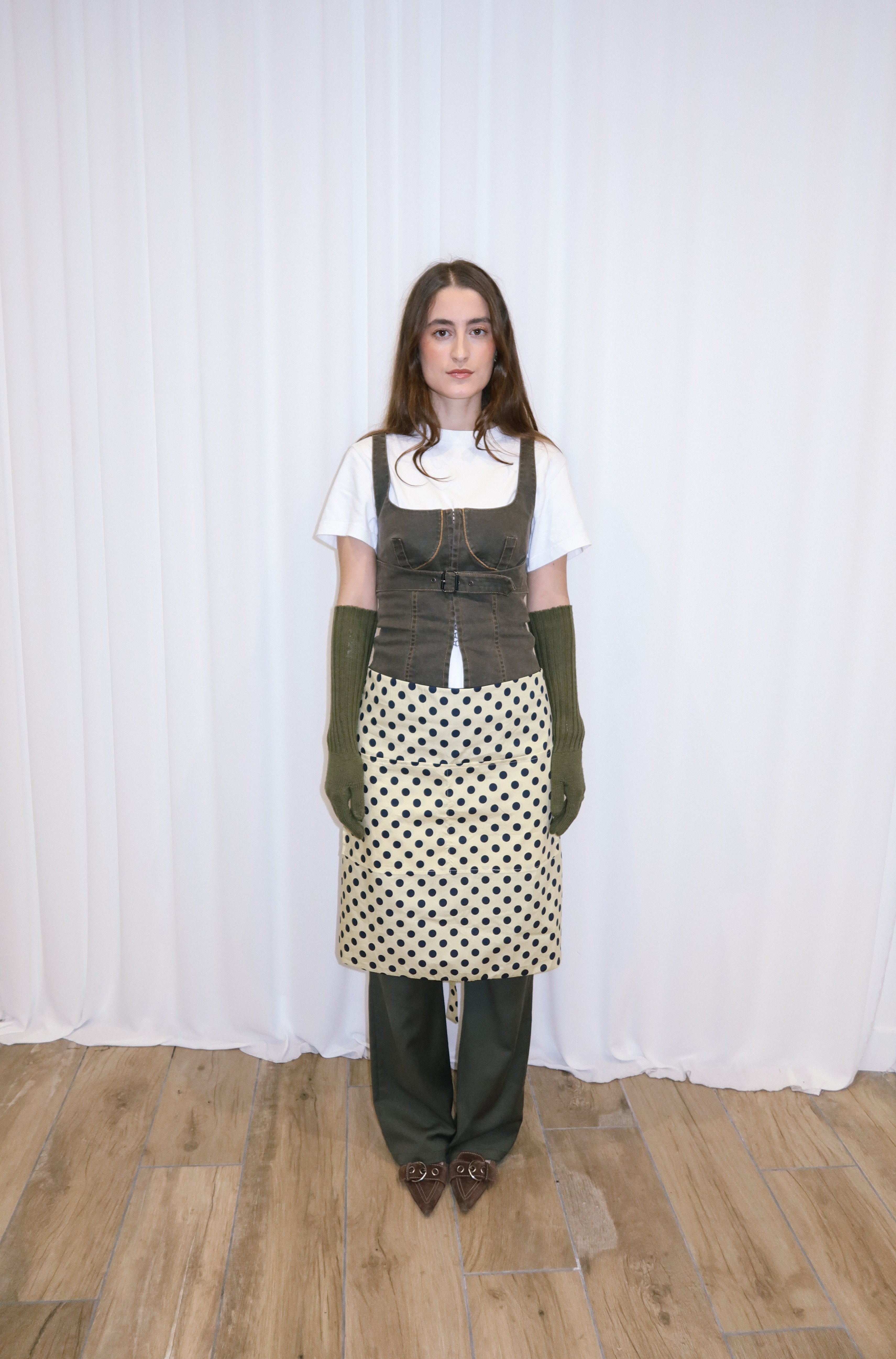 Person wearing a white shirt, polka dot apron, and green gloves against a white curtain background.