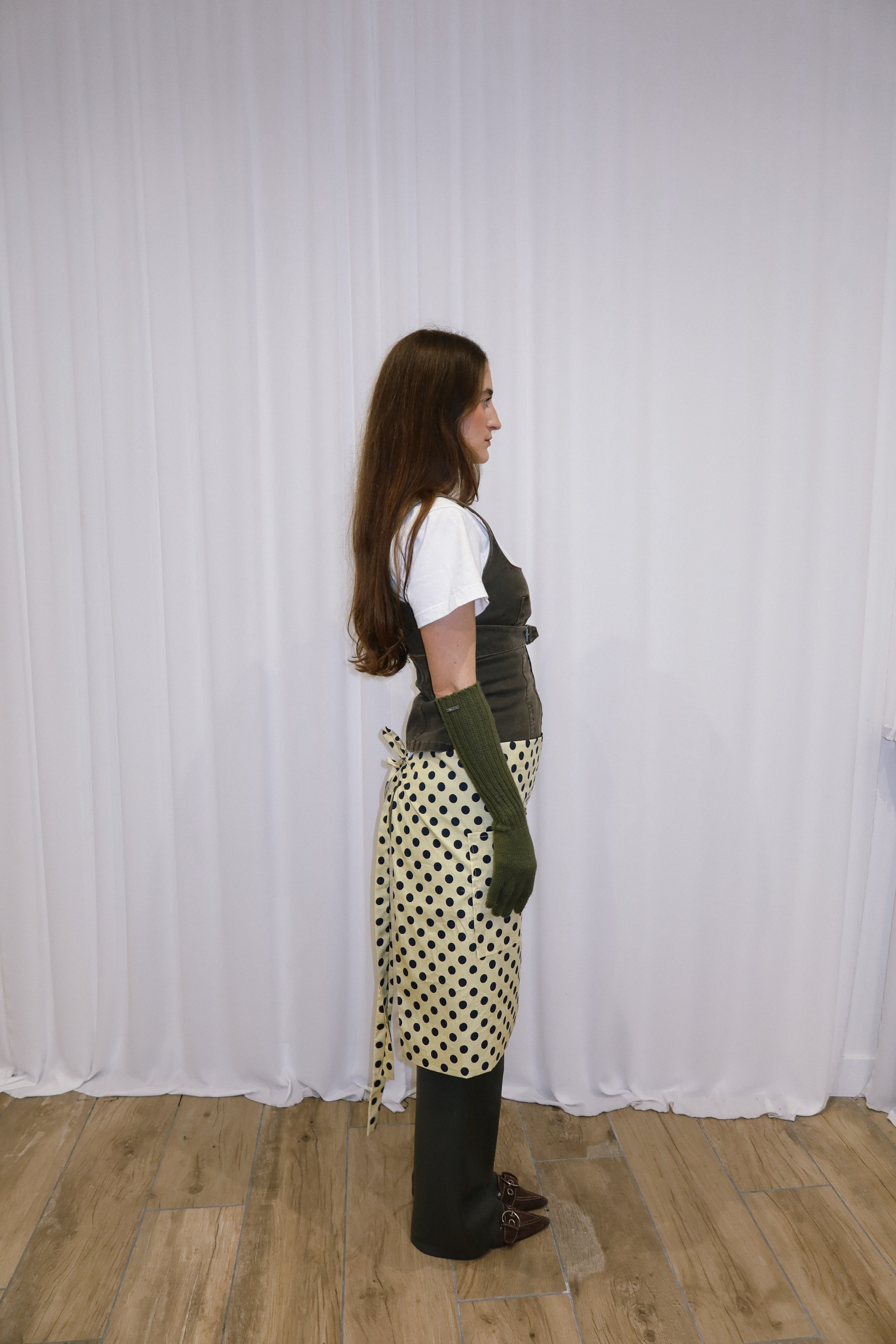Person wearing a white shirt, polka dot apron, and green gloves against a white curtain background.