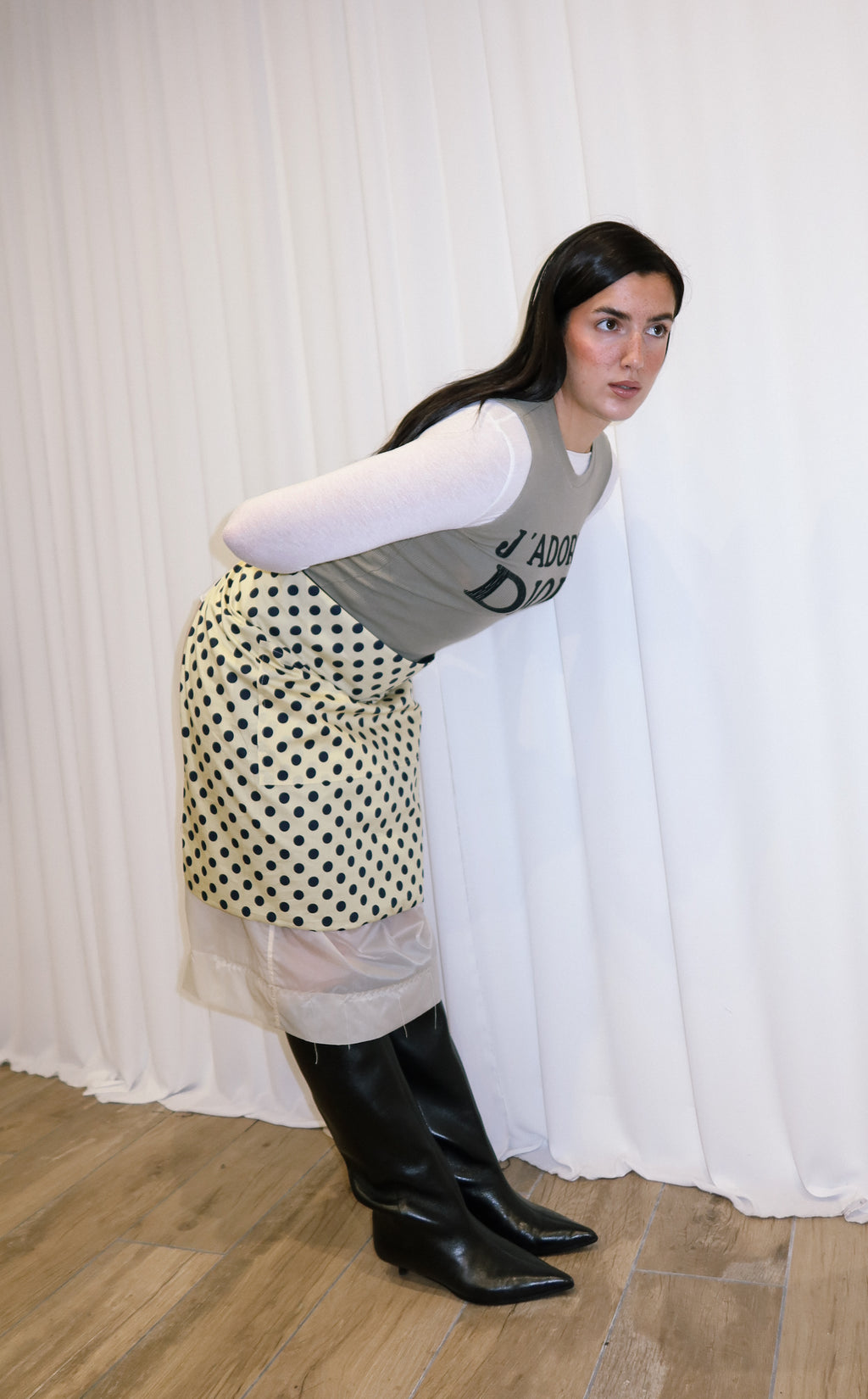 Woman wearing a patterned apron, gray top, and black boots against a white curtain background