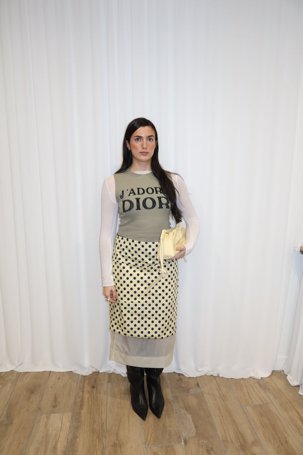 Woman wearing a patterned apron, gray top, and black boots against a white curtain background