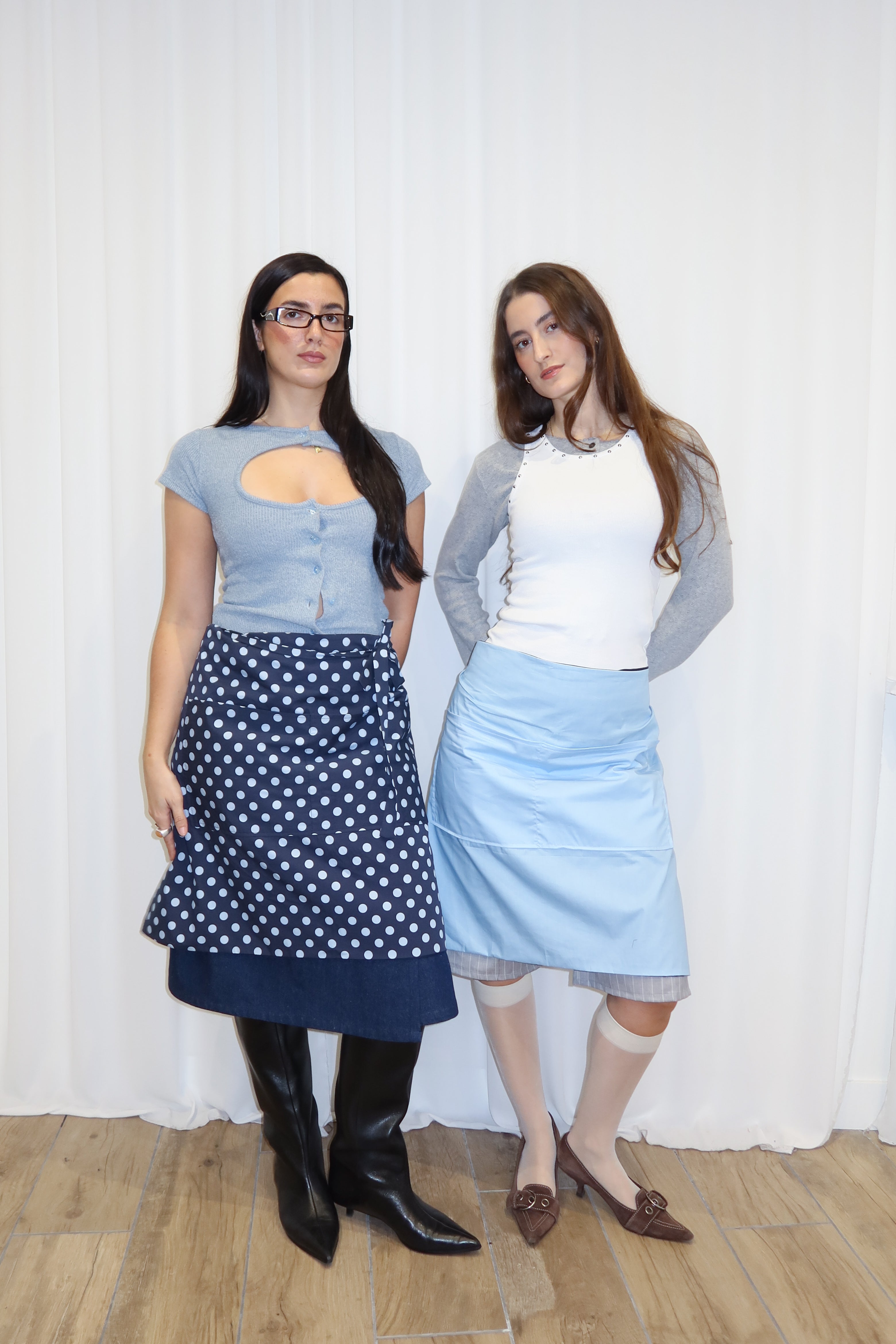 Two women standing side by side against a white curtain, wearing navy polka dots and light blue aprons.